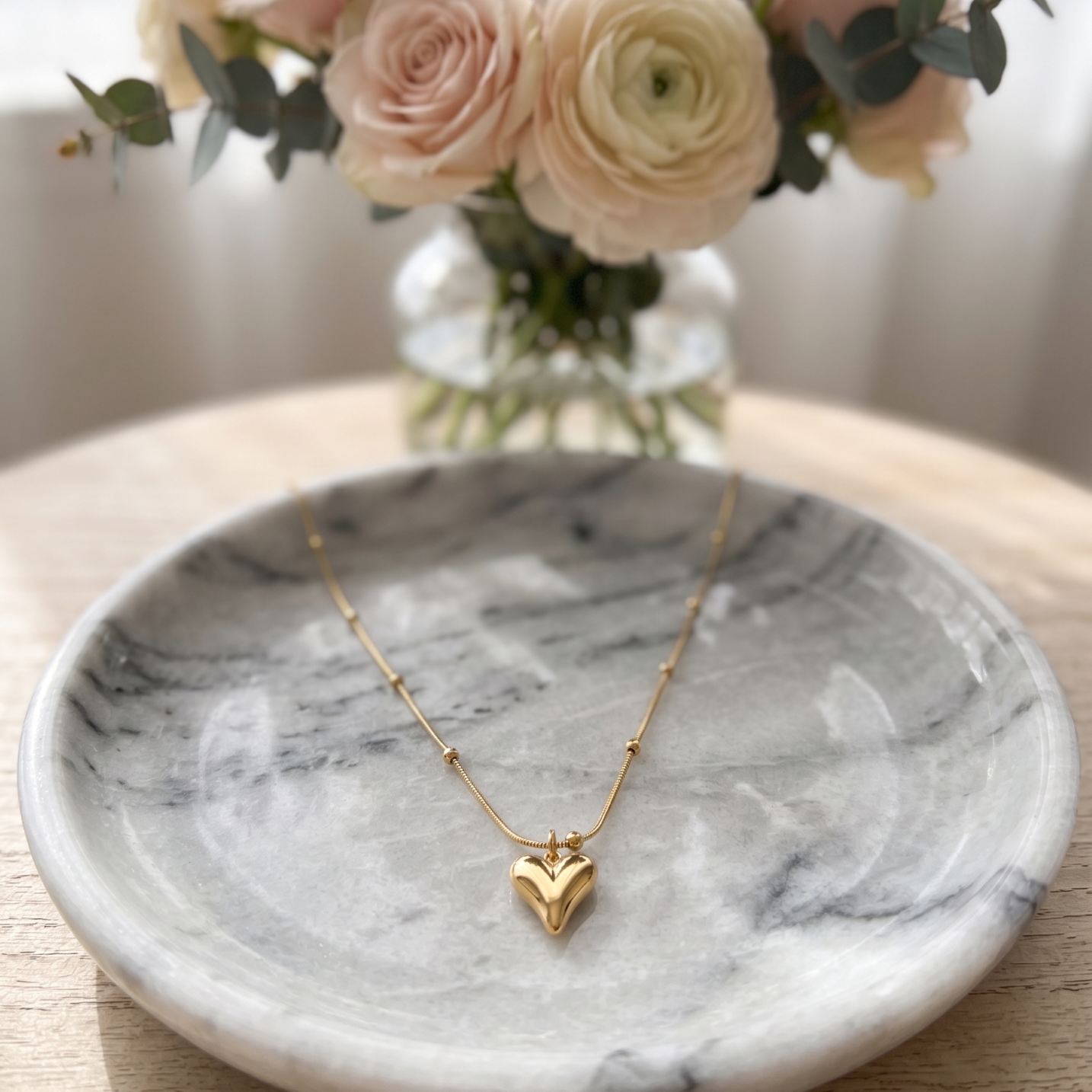 Gold necklace with a heart pendant on a marble dish, with a vase of flowers in the background.