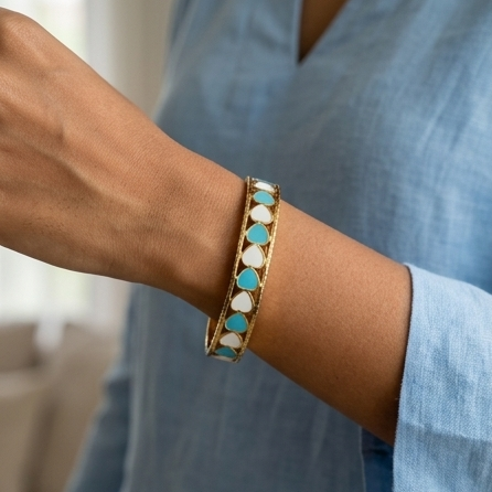 Close-up of a wrist wearing a gold bracelet with turquoise and white stones, against a blurred indoor background.