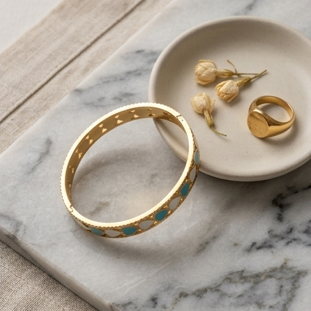 Gold bracelet with turquoise stones on a marble surface with a small dish containing dried flowers and another gold ring.